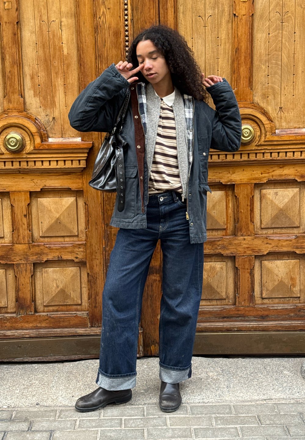 Femme aux cheveux bouclés portant des vestes superposées, une chemise rayée, un jean large, des bottes noires et un sac à main, posant les yeux fermés devant une porte en bois.
