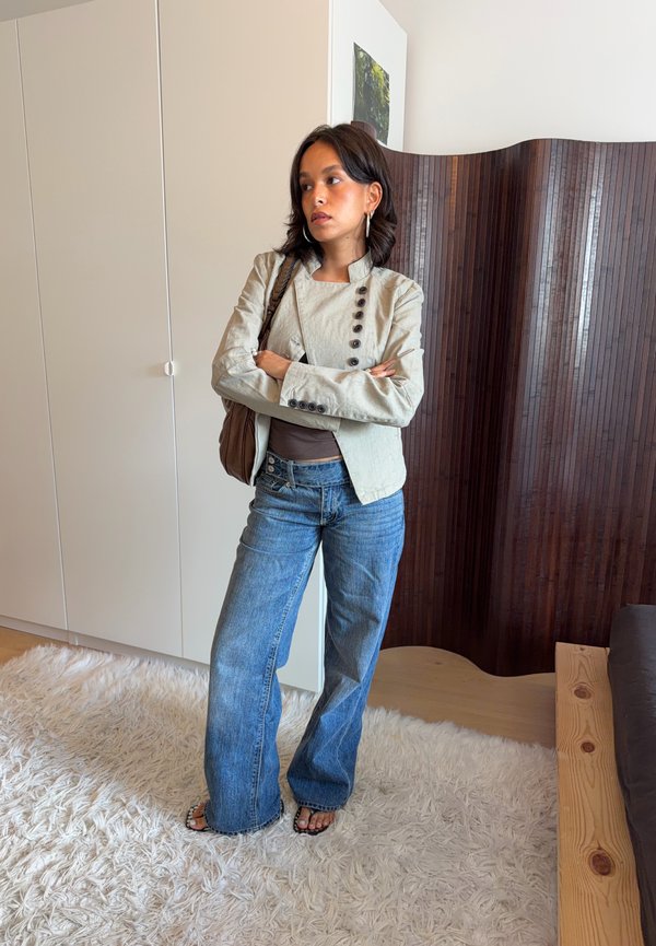 Mujer de pie con los brazos cruzados, vestida con una chaqueta beige, vaqueros azules y sandalias en una habitación con armario blanco y biombo de madera.