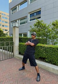 Camicetta nera a maniche corte, pantaloni corti abbinati e calzature nere senza lacci; indossa un cappello lavorato a maglia con motivo e occhiali da sole, posato all'aperto.