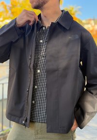 Άνδρας φοράει μαύρο μπουφάν North Face πάνω από καρό πουκάμισο με κουμπιά, με το ένα χέρι να ρυθμίζει το φερμουάρ του μπουφάν και το άλλο στην τσέπη.