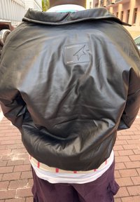 Black leather bomber jacket with a smooth texture, featuring a logo patch on the back. Worn over a white shirt and purple shorts.