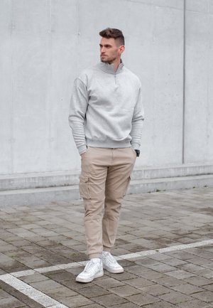 Hombre con pantalones cargo beige, sudadera gris con cremallera corta y zapatillas blancas, de pie sobre suelo pavimentado frente a una pared de concreto gris.