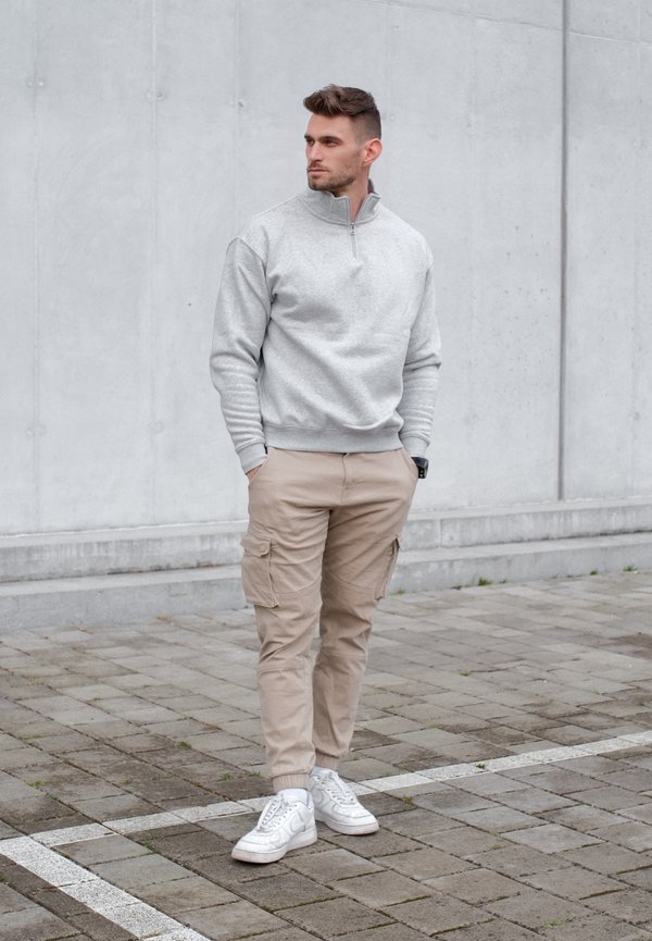 Homme en pantalon cargo beige, sweat-shirt gris à demi-fermeture éclair et baskets blanches, debout sur un sol pavé devant un mur en béton gris.