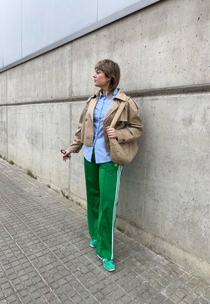 Un abrigo trinchera recortado beige sobre una camisa azul claro, combinado con pantalones deportivos verdes con franjas blancas y zapatillas a juego.