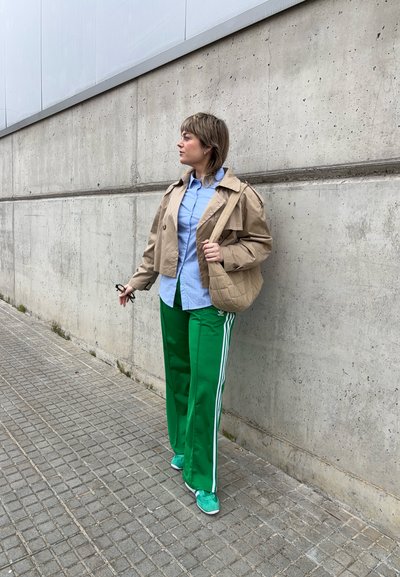 Un abrigo trinchera recortado beige sobre una camisa azul claro, combinado con pantalones deportivos verdes con franjas blancas y zapatillas a juego.