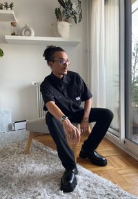 Zwarte T-shirt met korte mouwen en twee borstzakken, zwarte broek en dikke zwarte schoenen. Zit op een lichtgekleurde stoel met een tapijt.