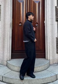 Homme en veste et pantalon noirs debout sur des marches en pierre courbes devant une grande porte en bois avec des poignées circulaires.