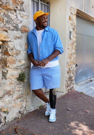 Camisa de botones azul claro, camiseta blanca, pantalones cortos a rayas azules y blancas, y zapatillas deportivas. Pierna prostética negra. Gorra amarilla y gafas de sol redondas.