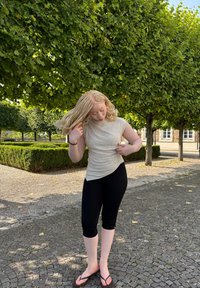 Top curto de manga curta em bege claro com design drapeado, combinado com leggings capri pretas e chinelos em um caminho de paralelepípedos rodeado de vegetação.
