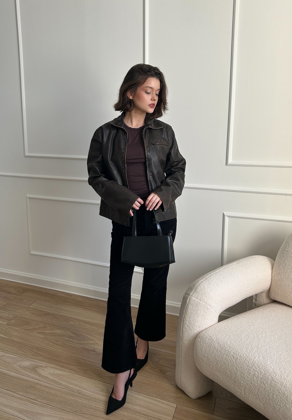 Barna bőrkabát egy testhezálló bordó toppal, fekete trapéznadrággal és fekete táskával. A színen világos fa padló és egy krém színű kanapé található.