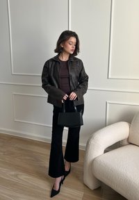 Barna bőrkabát egy testhezálló bordó toppal, fekete trapéznadrággal és fekete táskával. A színen világos fa padló és egy krém színű kanapé található.