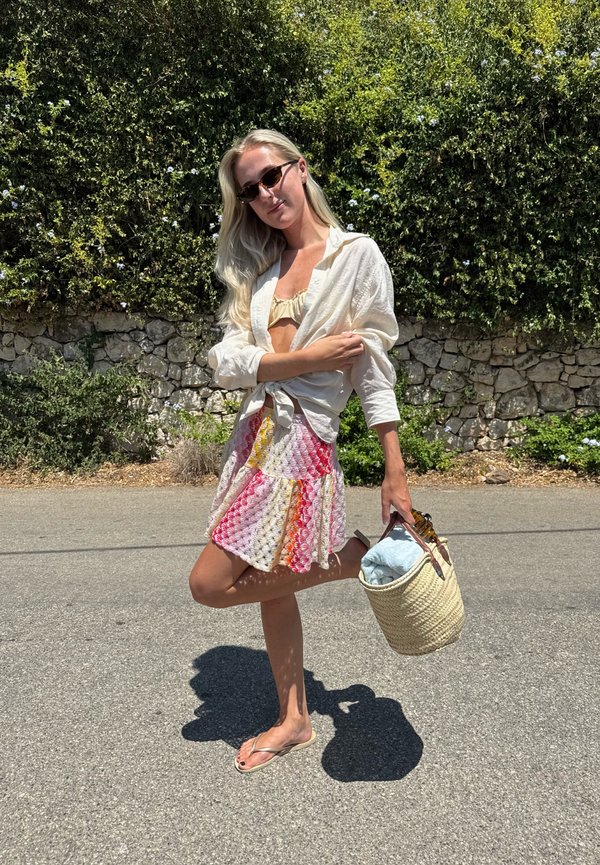 Femme portant des lunettes de soleil, une chemise blanche, une jupe colorée et des tongs tient un sac en osier et pose sur une route ensoleillée près d'un mur de pierre et de la verdure.