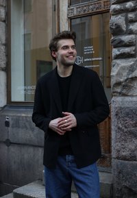 Blazer preto sobre um suéter preto, combinado com jeans azuis. De pé do lado de fora de um edifício, encostado a uma parede de pedra texturizada, mostrando uma porta com vidro.
