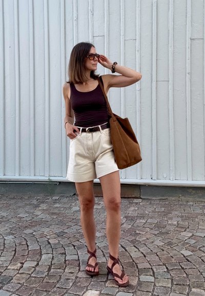 Bolso tote marrón, camiseta sin mangas negra, pantalones cortos color crema, sandalias marrones. De pie al aire libre con una superficie de piedra texturizada y un fondo de madera vertical.