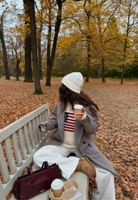 Manteau à motif pied-de-poule gris, bonnet en maille torsadée blanc, pull crème avec des rayures rouges et bleues. Sac à main bordeaux, pantalon blanc, tasse de café à la main.