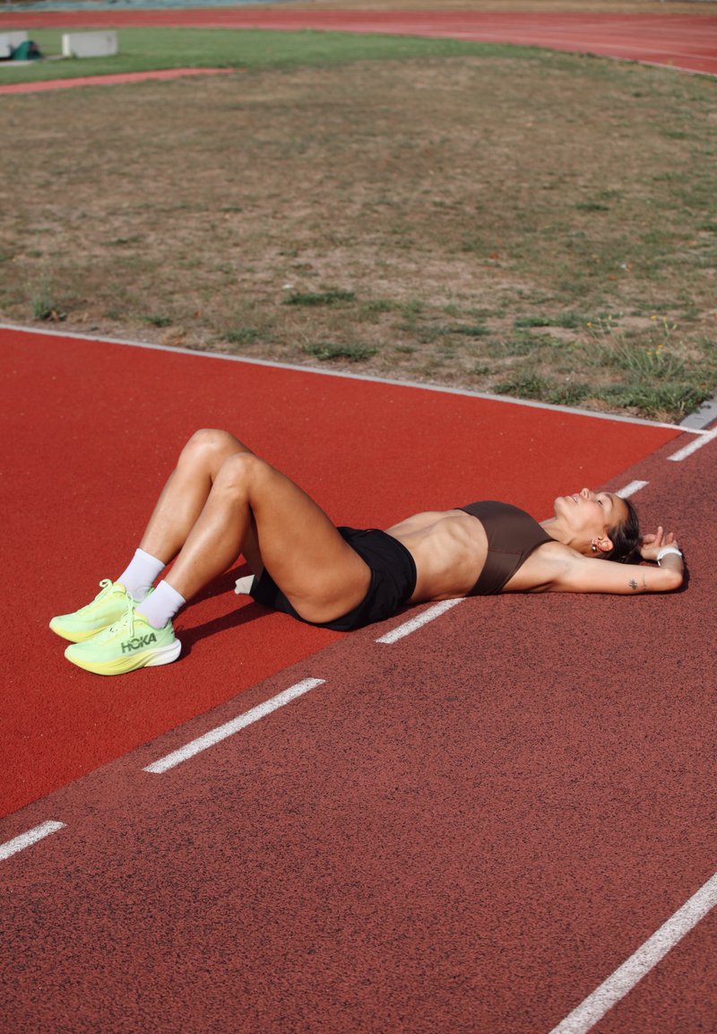Donna in abbigliamento sportivo sdraiata sulla schiena su una pista di atletica rossa con le braccia distese dietro la testa, che riposa dopo l'esercizio.
