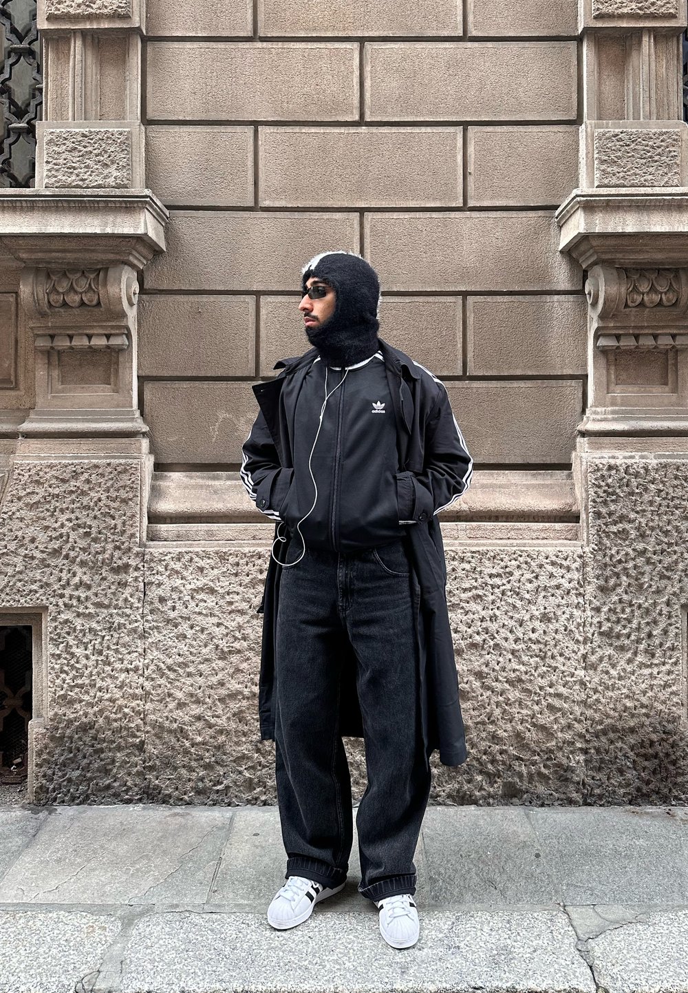 Homme portant un manteau noir, une veste Adidas, un cagoule, un jean foncé et des baskets blanches, debout contre un mur en pierre texturé, les mains dans les poches, regardant sur le côté.