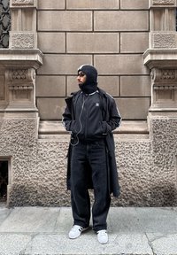 Homme portant un manteau noir, une veste Adidas, un cagoule, un jean foncé et des baskets blanches, debout contre un mur en pierre texturé, les mains dans les poches, regardant sur le côté.