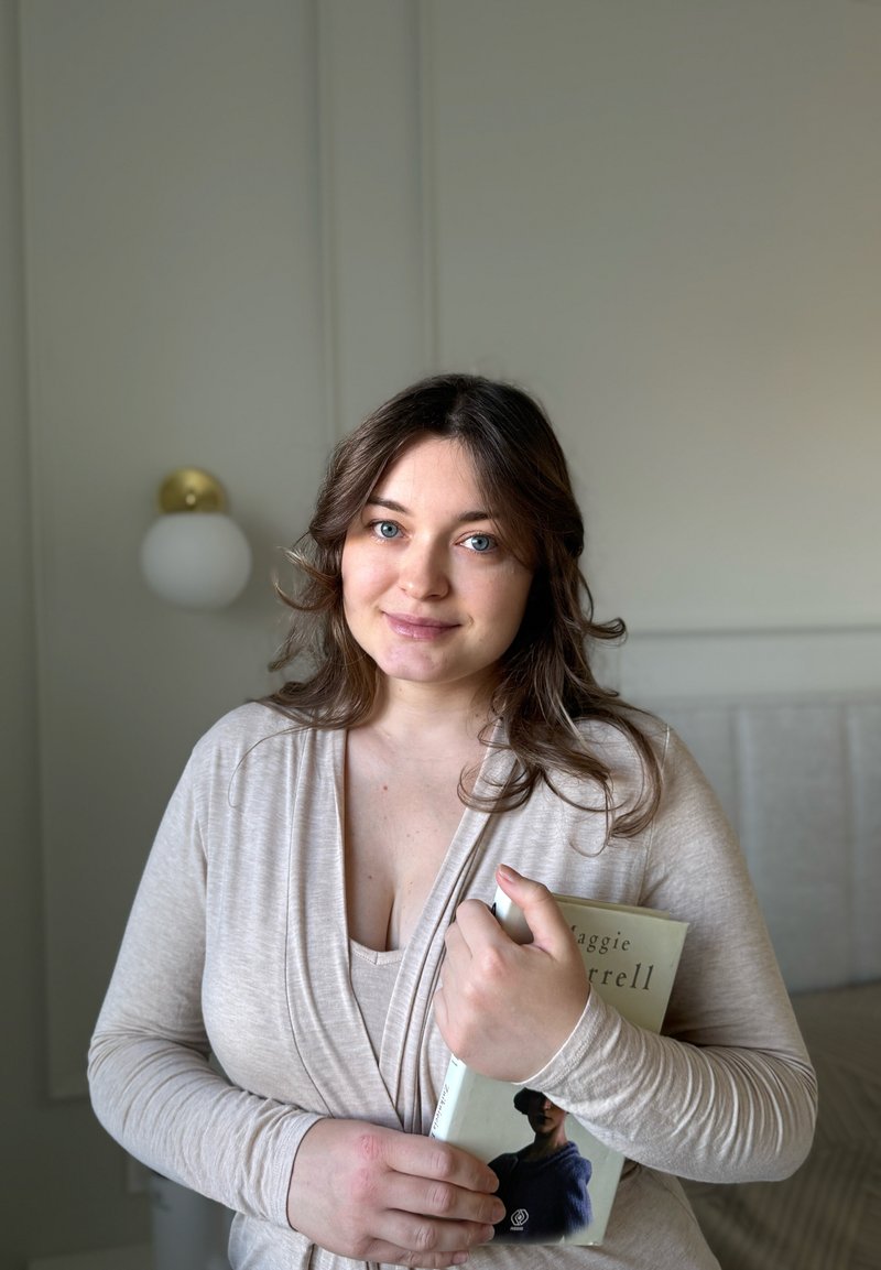 Haut beige clair à manches longues avec encolure drapée porté par une personne tenant un livre. Fond texturé avec lumière douce et lampe ronde.