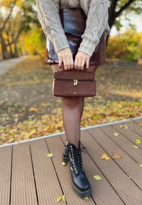 Brauner Lackleder-Minirock, beiger Zopfmuster-Pullover, schwarze Plateau-Stiefeletten und braune Wildleder-Clutch mit einem goldenen Reißverschlussdetails.