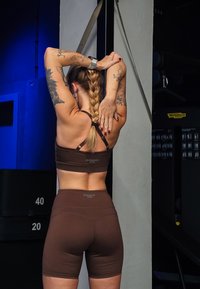 Ensemble de vêtements de sport brun comprenant un crop top et un short taille haute. Le matériau semble extensible avec une texture lisse. Logo de marque notable affiché.