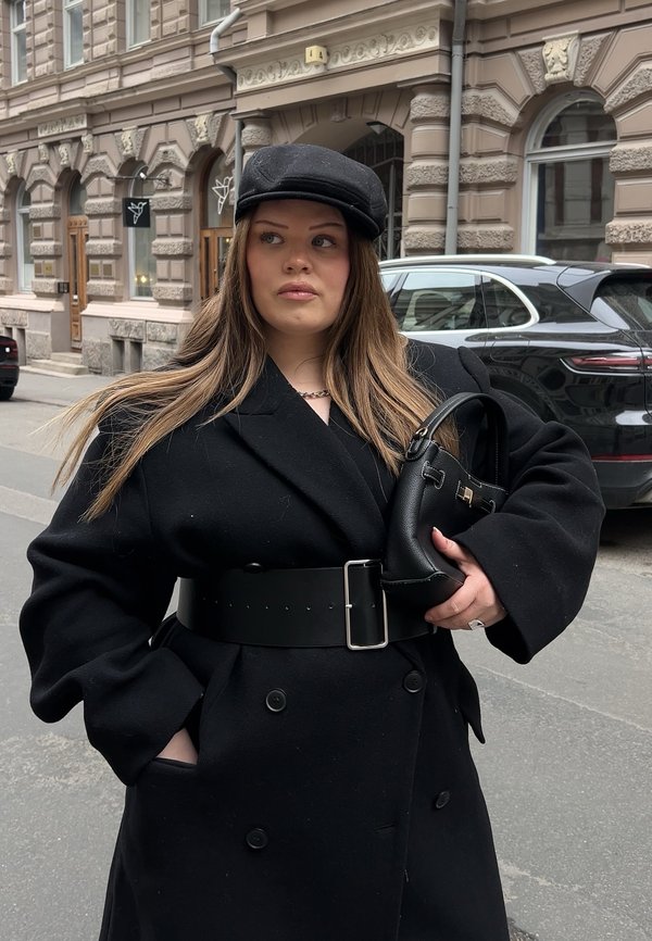 Femme aux longs cheveux portant un manteau noir, une large ceinture et une casquette noire, tenant un petit sac noir dans une rue de la ville avec des voitures garées.