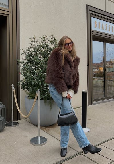 Chaqueta de piel sintética marrón, suéter de punto ligero, jeans azules, bolso negro, botas de tacón cuadrado negro, entorno al aire libre con vegetación y un edificio.