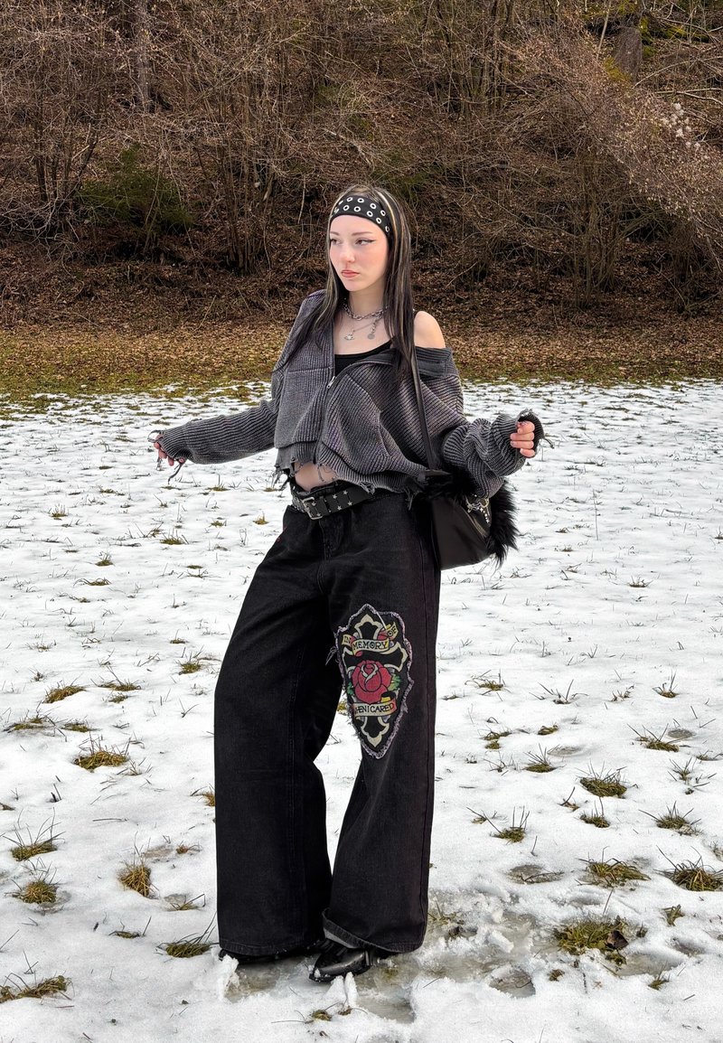 Jeune femme aux longs cheveux bruns debout sur de l'herbe recouverte de neige, portant un pull court gris, un pantalon large noir avec un patch en forme de rose, et un bandeau.