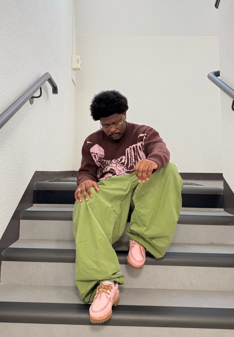 Maglione marrone con grafica rosa, pantaloni verdi a gamba larga e scarpe rosa con suole color nocciola, seduta sui gradini con un corrimano testurizzato.