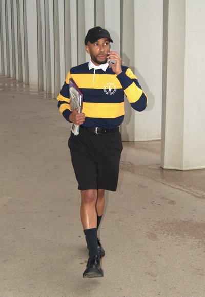 Camisa de polo a rayas azul y amarilla, pantalones cortos negros, gorra negra, sosteniendo carpetas, caminando sobre un suelo liso, con un fondo de columnas.