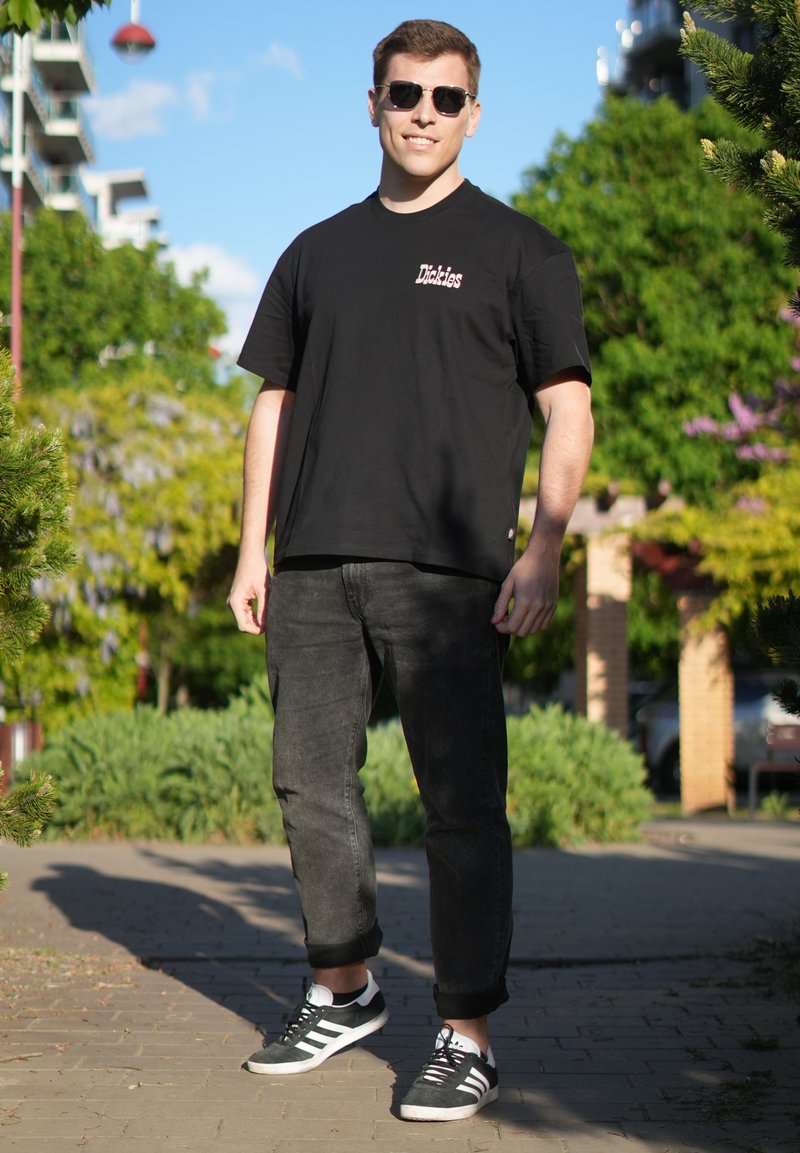 Jeune homme portant un t-shirt noir Dickies, un jean noir retroussé et des baskets noires et blanches, debout en extérieur par une journée ensoleillée.