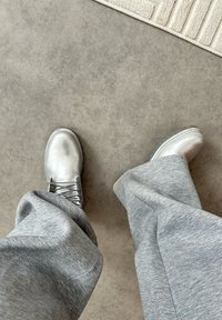 Person iført sølvfarvede metalliske snørestøvler og løstsiddende grå bukser, stående på en grå gulvbelægning med kanten af et beige tæppe synlig.