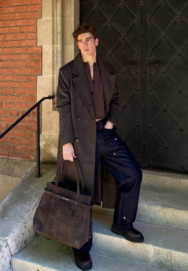 Black oversized coat, dark brown top, deep blue jeans, and black footwear. Carrying a dark brown leather tote bag with sturdy handles.