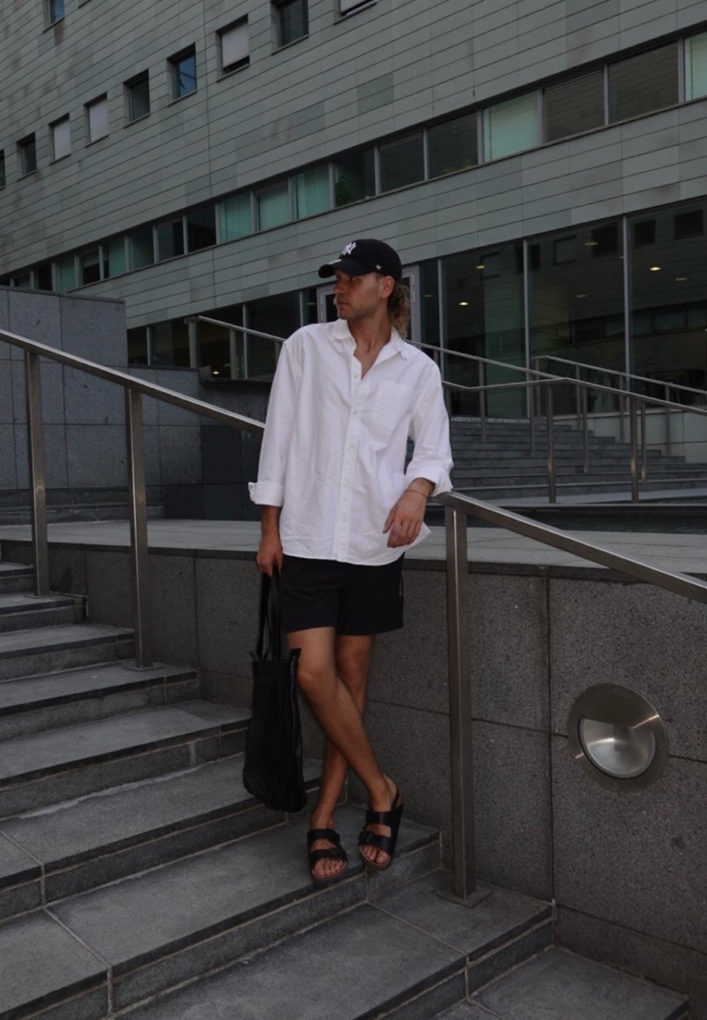 Hvit knappopp skjorte, svarte shorts, svarte flip-flops og svart tote bag; står på steintrinn ved siden av et metallrekkverk og en modern bygning.