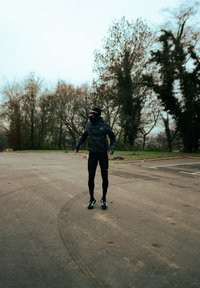 Athlète portant une veste noire texturée et des leggings noirs moulants se tient à l'extérieur. Fond gris avec des arbres ; surface en asphalte montrant des traces de pneus.