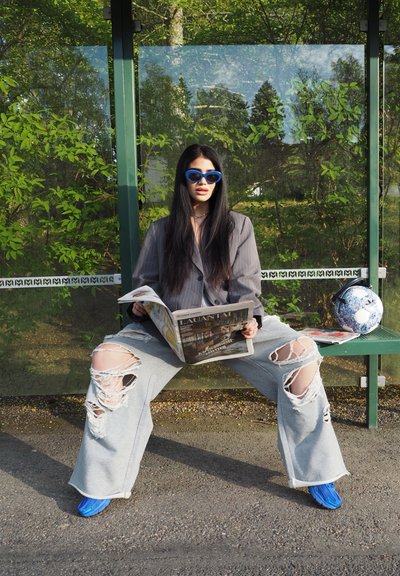Pantalones gris grandes con rodillas desgastadas, blazer negro y gafas de sol azules; sentado en un banco verde, leyendo un periódico.