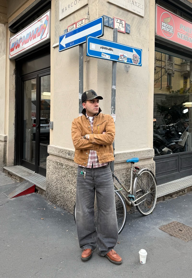 Homme en veste marron et casquette, les bras croisés, debout près d'un vélo, à côté de panneaux de rue indiquant à gauche et à droite. Gobelet de café posé sur le sol à proximité.