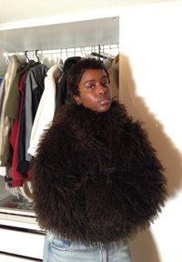 Vêtement d'extérieur en fausse fourrure marron avec un design volumineux et texturé, porté avec un jean bleu, sur fond de vêtements suspendus.