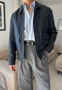 Casaco de couro azul-marinho com fecho éclair, dois bolsos frontais, combinado com uma camisa de botão azul-clara e calças cinza plissadas.