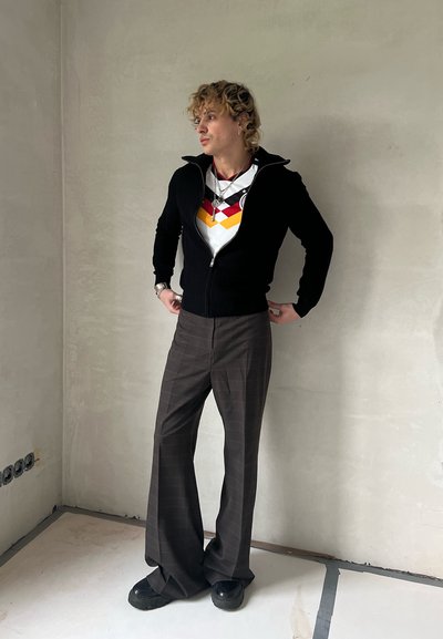 Suéter negro con cremallera, camisa blanca con patrón geométrico en rojo y amarillo, pantalones acampanados a cuadros marrones y zapatos negros. Fondo neutro.