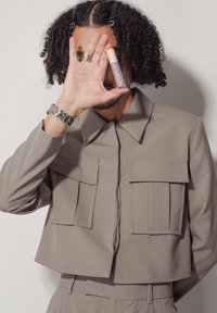 Veste beige courte avec de grandes poches avant et un col pointu. Tient un tube cosmétique lavande avec un capuchon beige clair.