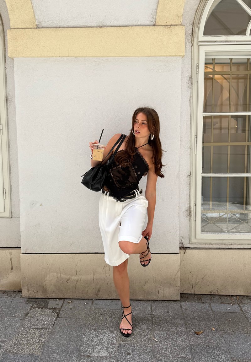 Jeune femme en haut noir et short blanc ajuste la lanière de sa sandale tout en tenant un café glacé et un sac à main noir près d’un mur blanc et d’une fenêtre.