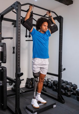Hombre con pierna prostética realizando una dominada asistida con eslingas en un equipo de gimnasio, de pie sobre una plataforma en una sala de fitness.