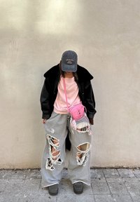 Personne portant une veste noire, une chemise rose, un pantalon large gris déchiré, une casquette grise et un sac bandoulière rose, debout contre un mur beige.