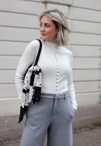 Femme aux cheveux blonds mi-longs portant un pull blanc à boutons et un pantalon gris, portant un sac noir avec un foulard à pois en plein air.