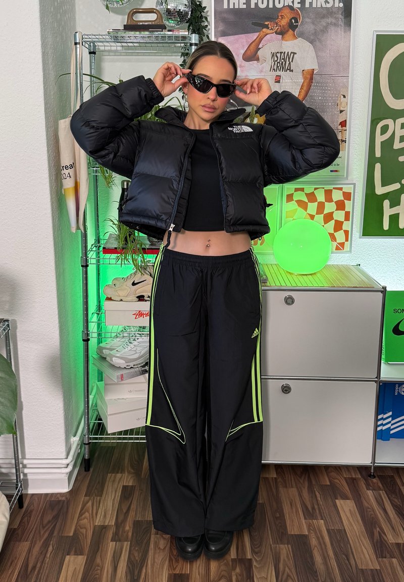 Black puffer jacket, cropped, with logo; black top; black pants with neon green accents and stripes; styled with black sunglasses and chunky shoes.