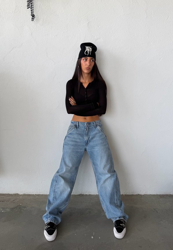 Kvinne iført svart langermet crop-top, lue med hvitt mønster, løse blå jeans og svarte joggesko står mot en hvit vegg med armene i kors.