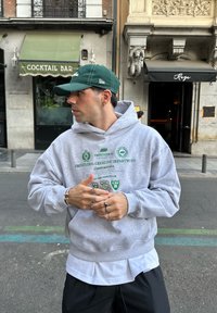 Grå hettegenser med grønn grafisk tekst, lagd over en hvit skjorte, brukt med svarte bukser og grønn baseballcap, synlige sølvringer.