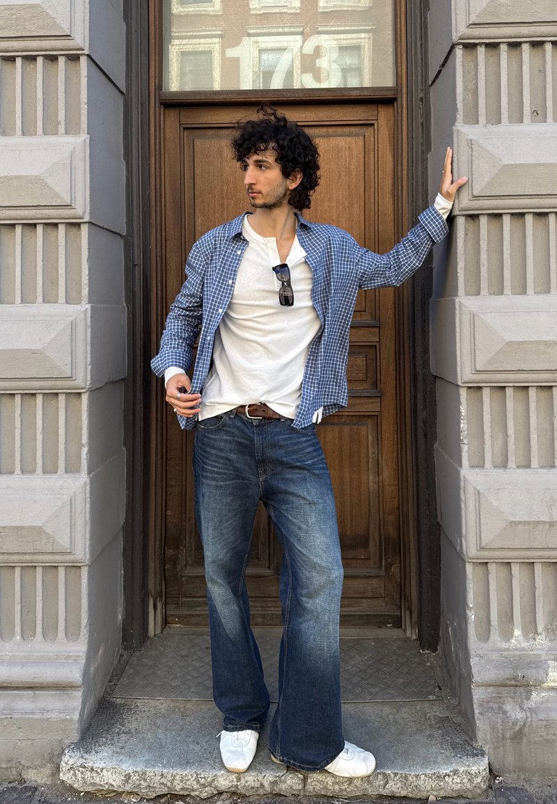 Homme aux cheveux bouclés, chemise à carreaux bleue, t-shirt blanc, jean évasé et chaussures blanches, debout sur un seuil de porte, bras écartés, regardant à gauche.