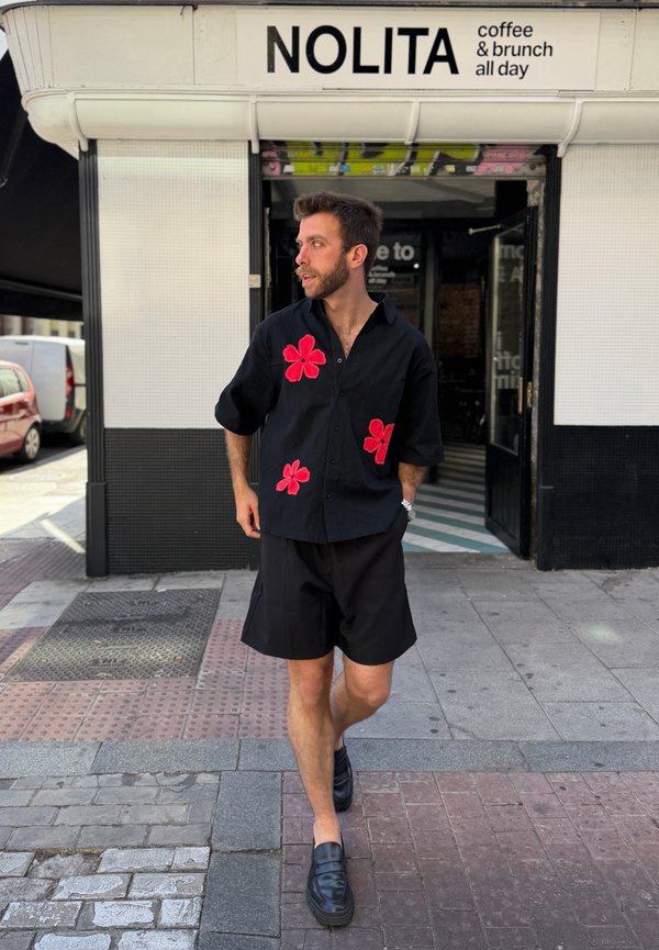 Homme portant une chemise noire à fleurs rouges et un short noir marchant devant un café nommé Nolita proposant café et brunch toute la journée.